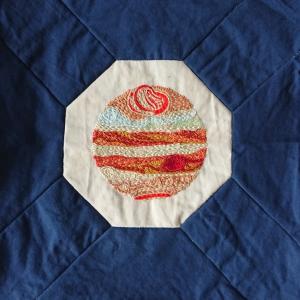 The planet Jupiter embroidered in white, yellow, orange and red threads on a white background, framed by blue fabric.