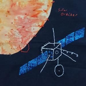 The solar orbiter satellite embroidered in white, with blue appliqued fabric to represent the solar panels. The sun appears in the top left corner in yellowy-orangey coloured fabric. The background is black. Red text is embroidered in the top right corner, it reads: Solar Orbiter.
