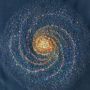 Spiral galaxy NGC 1232 created in different colour threads, predominantly orange, yellow, white and grey. In this version of the galaxy the spirals are in going in the opposite to the real life galaxy.