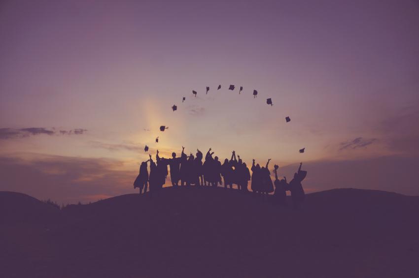 At sunset, the silhouette of a group or approximately 20 students standing on a hill throwing their mortarboard graduation caps in the air. Celebrating