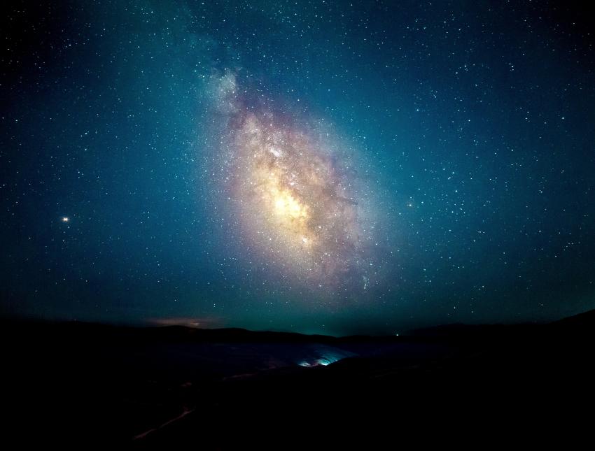 A photograph of the Milky Way over a mainly dark horizon with a valley illuminated by artificial light.