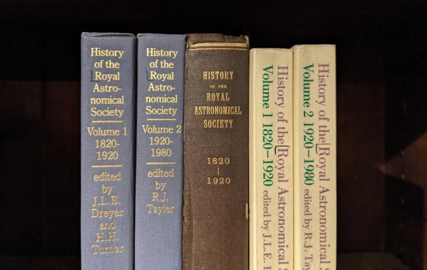Various editions (hardback, with gilt inlaid type, and paperback) of the History of the Royal Astronomical Society arranged vertically on a shelf.