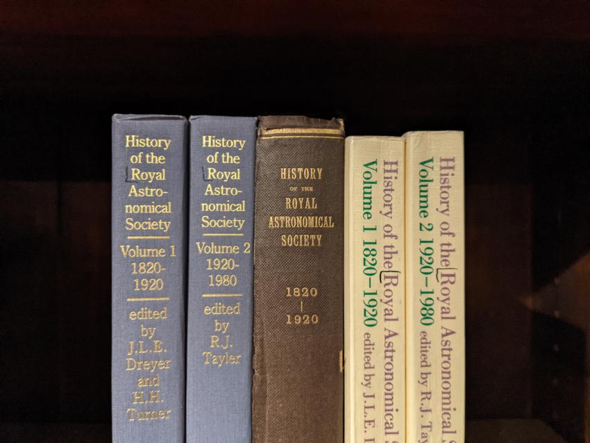 Various editions (hardback, with gilt inlaid type, and paperback) of the History of the Royal Astronomical Society arranged vertically on a shelf.