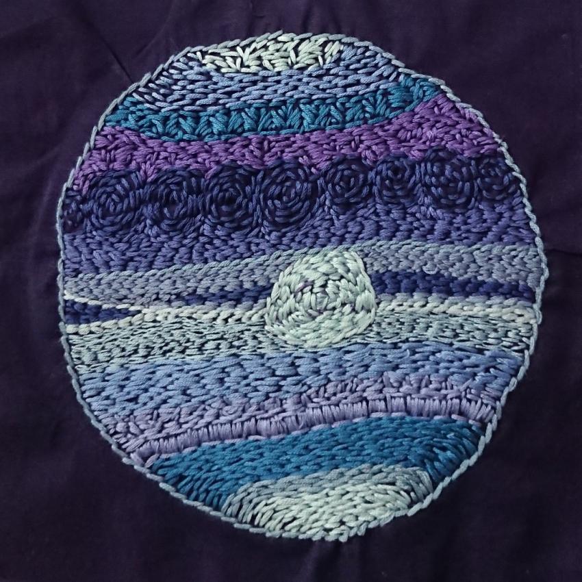 An imaginary planet, embroidery-thread doodled in shade of blue and purple coloured threads on a black background.