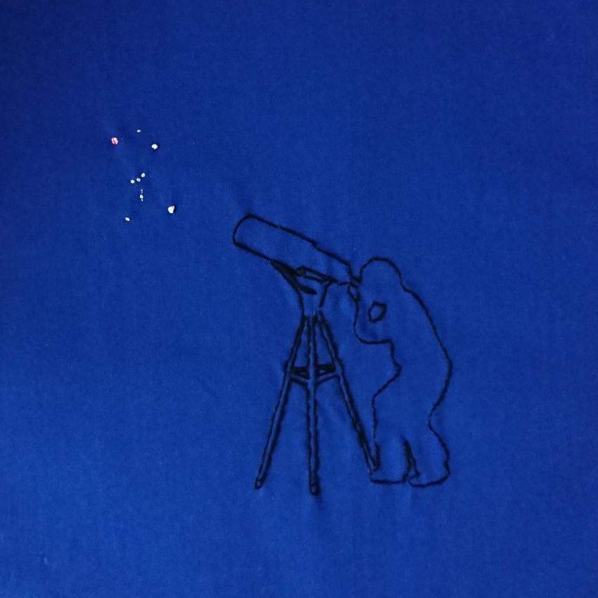 The outline of a person observing Orion through a telescope embroidered with black thread. The constellation of Orion is embroidered in white thread.