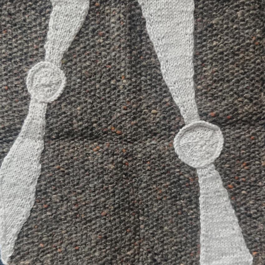 The square shows two pulsars, knitted in white wool on a grey wool background. Pulsars are spinning neutron stars that emit radio light in beams, which makes a lighthouse like effect (the pulsars are shown by the circles and the beams by the wedges coming from them). 