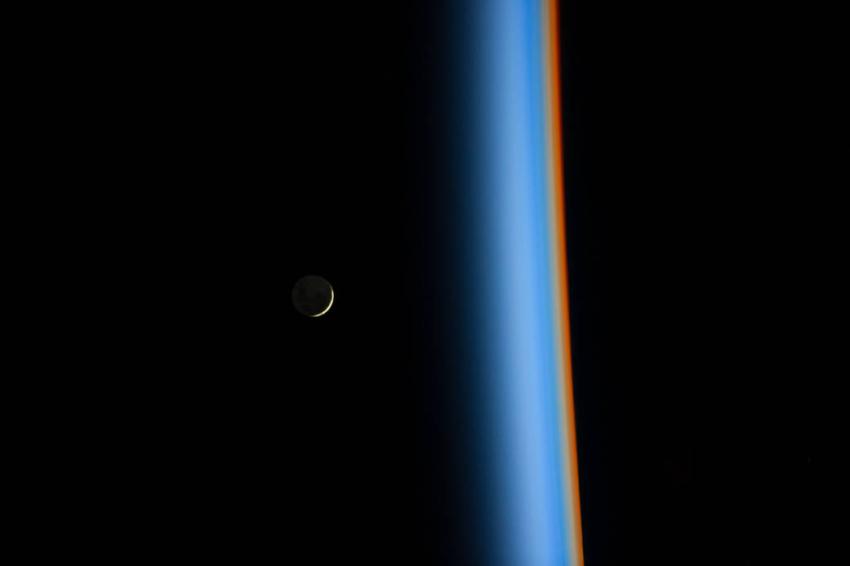 A view from the International Space Station of the Moon at left rising over the colourful bands of the Earth's atmosphere on the right.
