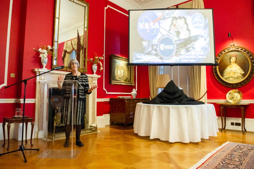 Professor Gillian Wright stands at a podium speaking to an audience. A screen behind her shows a graphic representing the MIRI instrument on the James Webb Space Telescope.