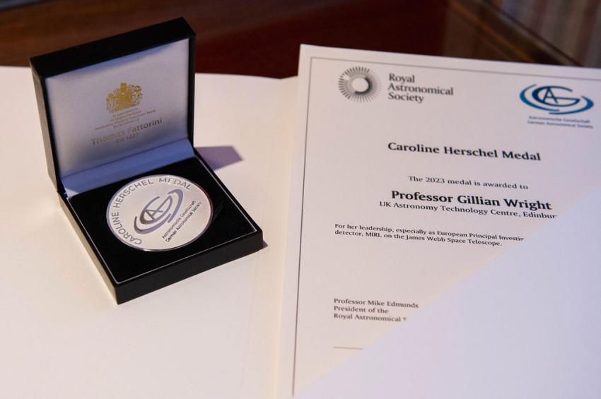 A silver medal in a black box is on a table next to a white certificate. The certificate bears the logos of the Royal Astronomical Society and the German Astronomical Society. Text on the certificate reads "Caroline Herschel Medal. The 2023 medal is awarded to Professor Gillian Wright for her leadership, especially as European Principle Investigator for MIRI on the James Webb Space Telescope." The certificate is signed by Professor Mike Edmunds and Professor Michael Kramer.