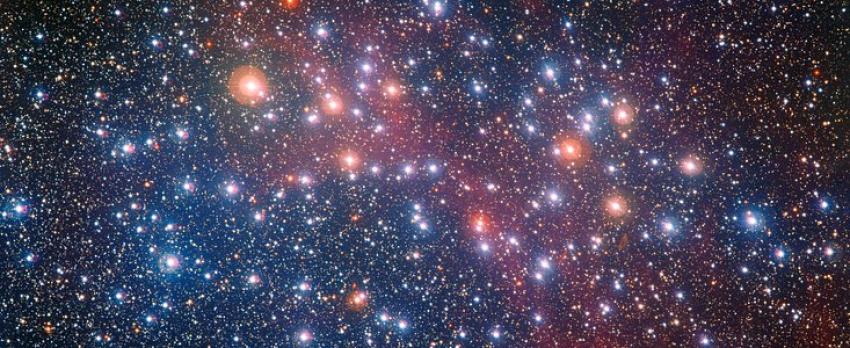 Image of the star cluster NGC 3532. Many bright stars of varying colours shine against a dark red and black backdrop of space.
