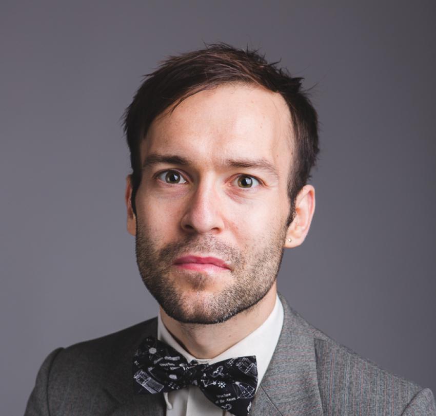 A headshot of the poet Dr Sam Illingworth wearing a grey coat and a bow tie with a white shirt.