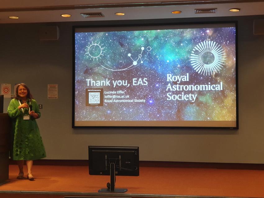 Woman in a green dress standing to the left on a stage with a colourful slide of gas and dust on the back wall. She just finished her talk and the slide says, "Thank you, EAS". 
