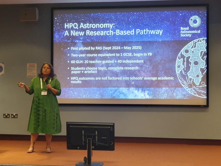 Woman in green dress standing on a stage presenting in front of a large screen behind her witha blue starfield and information on HPQ Astronomy. 