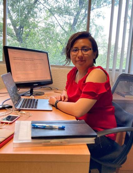 Image of Alejandra Lugo-Aranda at her work desk. A laptop is open in front of her, and another computer screen is visible in the background. She has dark short hair, wears glasses, and is wearing a red top. She sits in an office chair at her desk.