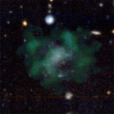 A diffuse galaxy in green and blue hues. White and orange stars appear around the clouds of the galaxy. 