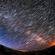 A wide angle view of the night sky and the Milky Way. Star trails appear across the entire sky, with several white lines (the satellites) overlapping the star trails.