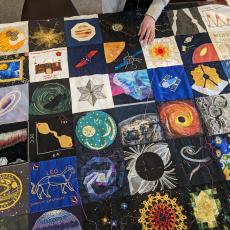 The bicentenary patchwork quilt laid out on a table, with a women working on stitching it together. It features embroidered squares depicting galaxies, planets and other celestial objects. 