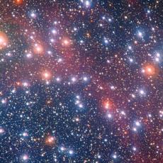 Image of the star cluster NGC 3532. Many bright stars of varying colours shine against a dark red and black backdrop of space.
