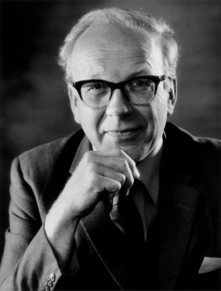 A black and white portrait of Professor Sir Francis Graham Smith. He rests his chin on his hand and smiles at the camera. He is wearing glasses, a shirt, tie and blazer.