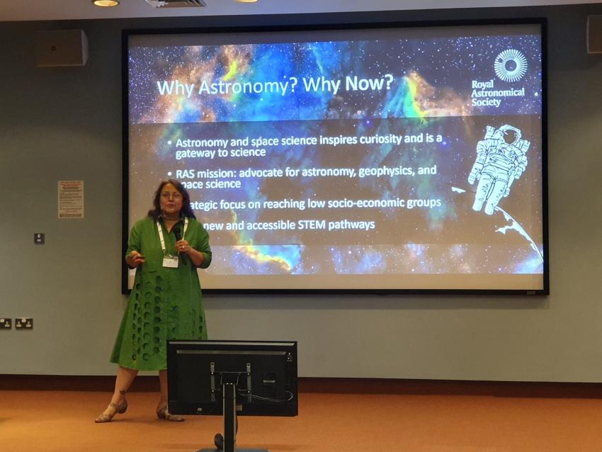Woman in green dress standing on a stage in front of a screen with a vibrant nebula on a slide describing why teach astronomy. 