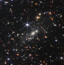 A large number of galaxies of different colours, shapes and sizes against the black backdrop of space. Galaxies in the middle of the image appear distorted due to gravitational lensing.